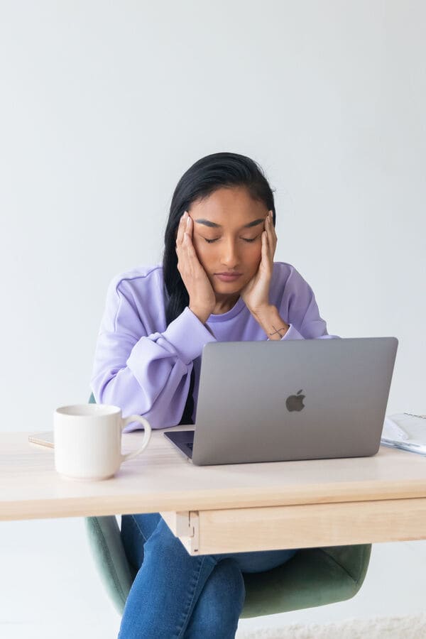 woman holding her head with her eyes closed, sitting in front of a laptop