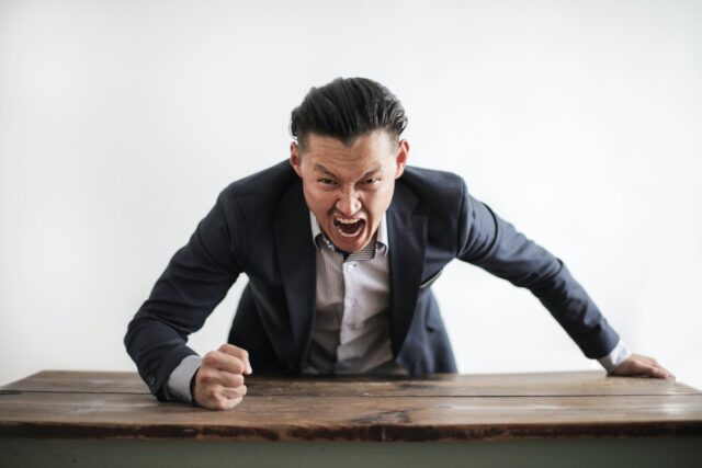 Expressive angry businessman in formal suit looking at camera and screaming with madness while hitting desk with fist